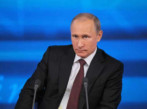 A serious-looking political figure seated at a press conference with a blue background